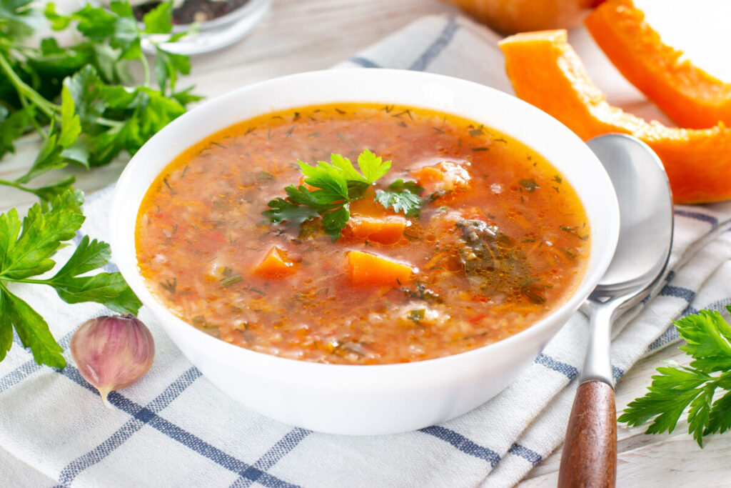 Овощной суп с кусочками тыквы и петрушкой в белой миске рядом с тыквой и чесноком.