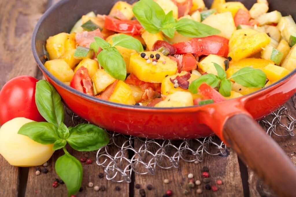 Картофельные кусочки с помидорами, кабачком и листьями базилика в красной посуде
