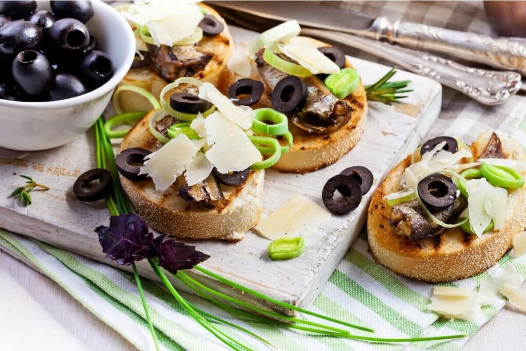 Небольшие тосты с рыбой, кольцами порея, оливками и сыром на белом блюде