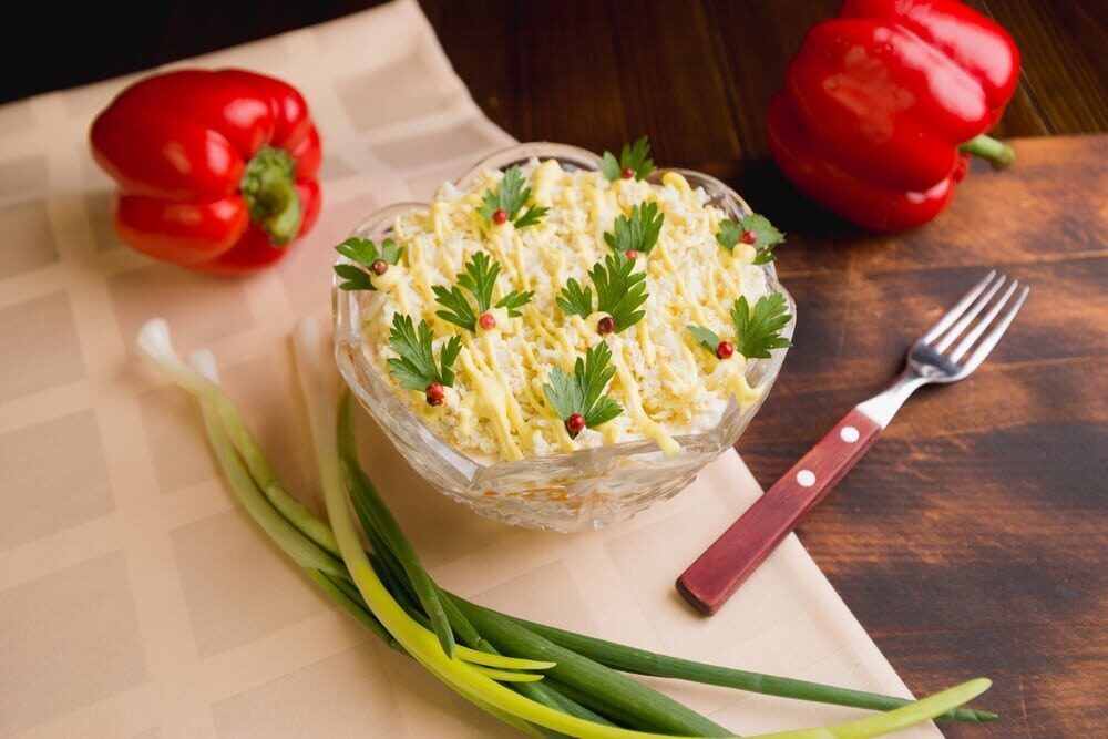 Слоёный салат с тёртыми яйцами и сыром в стеклянном салатнике, украшенный зеленью